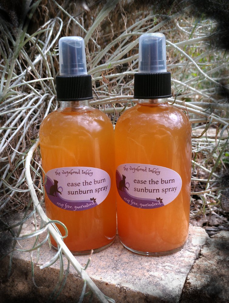 Two big clear glass spray bottles filled with golden-brown liquid. Each has a label that says 'ease the burn sunburn spray'.