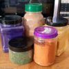Five jars, some glass, some plastic, holding a variety of brightly colored powders.