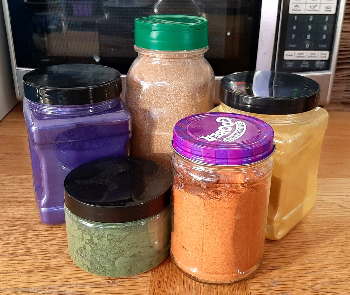 Five containers, some plastic, some glass, in varying sizes, sit on a table. They're filled with powders in varying colors -- rich blue, sparkly yellow, forest green, orange, and a light brown.
