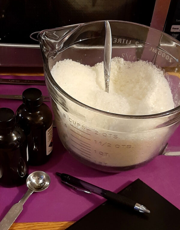 A big glass measuring cup is filled with white bath salts. Around it are a scattering of metal measuring spoons, two amber glass bottles, a notepad, and a pen.