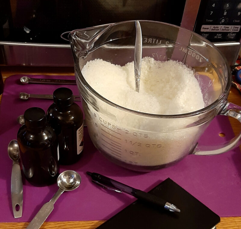 A big glass measuring cup is filled with white bath salts. Around it are a scattering of metal measuring spoons, two amber glass bottles, a notepad, and a pen.