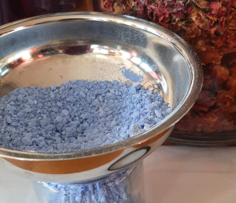 A wide metal funnel sits atop a clear glass jar; both are filled with pale blue bath salts.