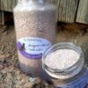Two clear glass jars filled with reddish-brown bath salts. One, the much shorter one, has been opened to show the bath salts inside.