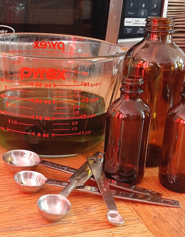 Three empty amber glass bottles sit next to a scattering of metal measuring spoons. Behind them is a big glass measuring bowl half-filled with yellow-green grapeseed oil.