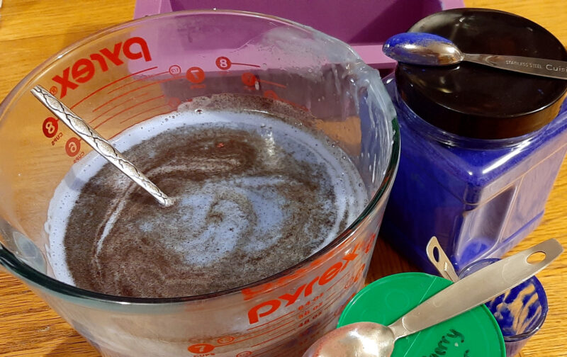 A big glass measuring bowl is filled with pale blue soap with a swirl of dark brown powder atop it. Sitting next to the bowl is a container of rich blue powder, a purple soap mold, and another container with a green lid.