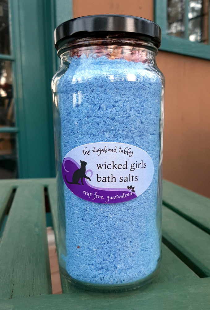 A tall clear glass jar filled with blue bath salts. The very top holds a layer of rose petals.