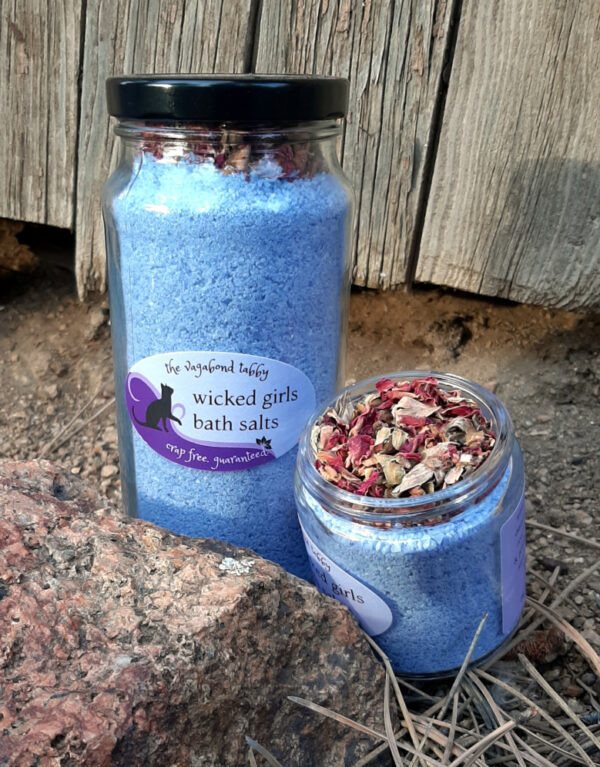 Two clear glass jars filled with blue bath salts. The very top of the taller one holds a layer of rose petals; the shorter one is open, and is also topped with a layer of rose petals.