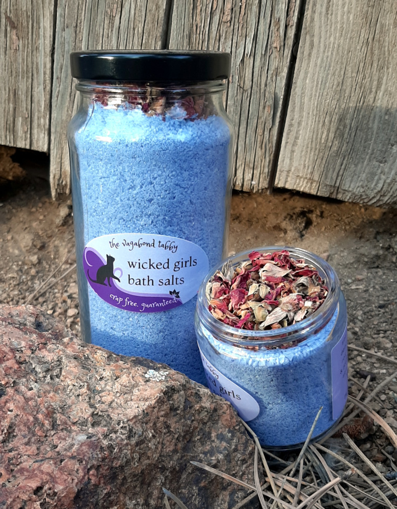 Two clear glass jars filled with blue bath salts. The very top of the taller one holds a layer of rose petals; the shorter one is open, and is also topped with a layer of rose petals.