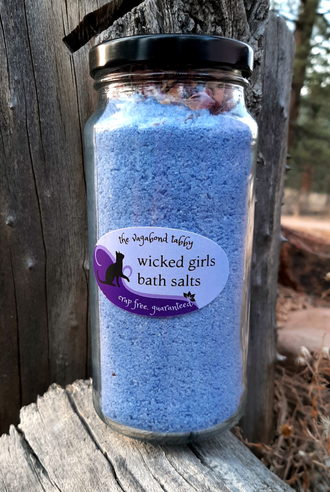 A tall clear glass jar filled with blue bath salts. The very top holds a layer of rose petals.