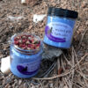 Two clear glass jars filled with blue bath salts. One is open, showing that the top of the jar is filled with rose petals.