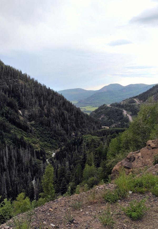 A precipitous mountain pass, with evergreen trees cloaking the sides & the glint of a rushing mountain stream at the bottom. In the distance are lower, more rounded hills against a bright but cloudy sky.