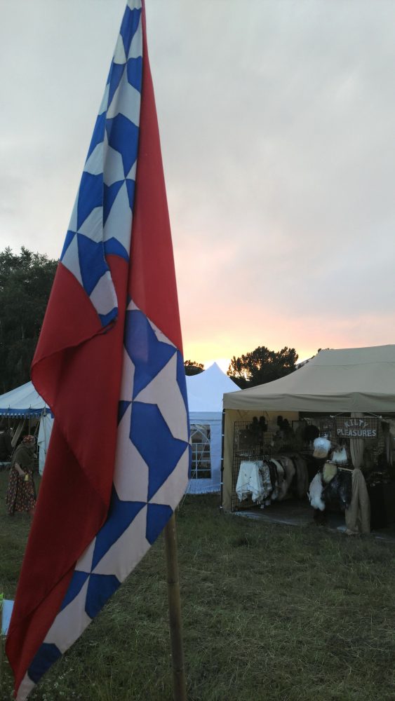 An SCA banner in red, white, & blue waves in front of the last vestiges of a yellow-fading-to-purple sunset.