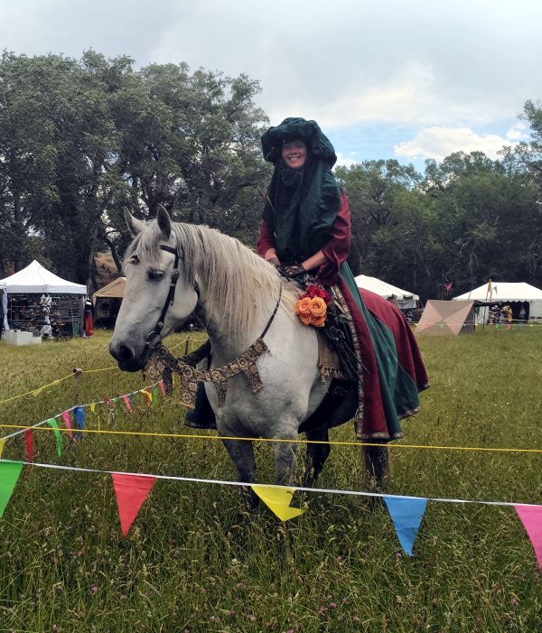 A white person in fancy medieval garbin deep red & green sits, smiling, atop a beautiful grey horse.