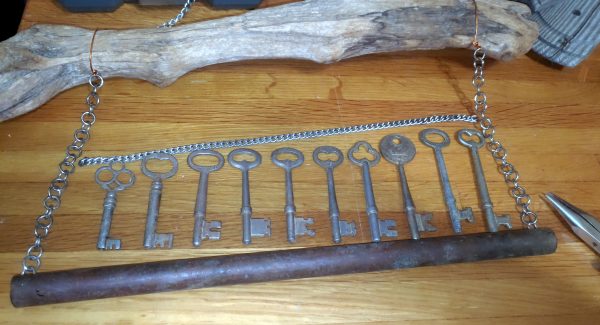 The stick and chime are laying on the work table. A short length of chain extends between the two chains holding the chime, and a bunch of skeleton keys, arranged shortest to longest, are lined up just below it.