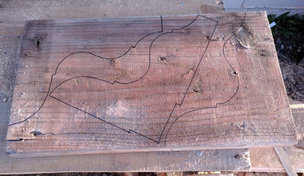 A large slab of wood sits on the worktable. The template's been traced onto it three times -- twice along the edges, but the third is at a funky angle right in the middle.