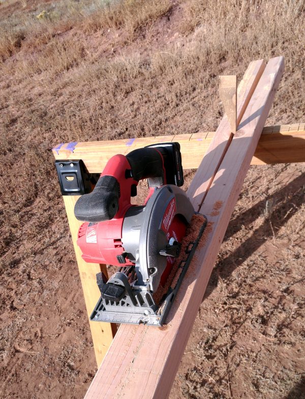 A 2x4 board, partway through being cut in half the long way. A circular saw sits in the middle of the board, & nehind it a wedge of wood holds the two already-cut parts separate.