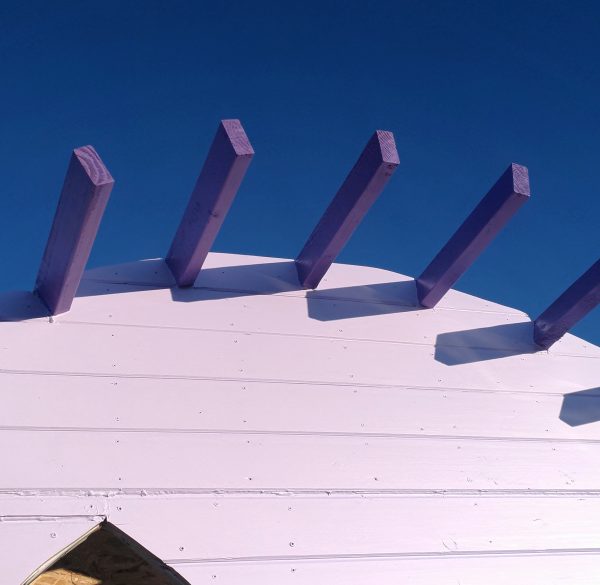A view looking upward at an angle -- the top of the front wall, painted lavender; the exposed parts of the rafters, painted a deeper purple; & behind it all, that deep blue New Mexico sky.