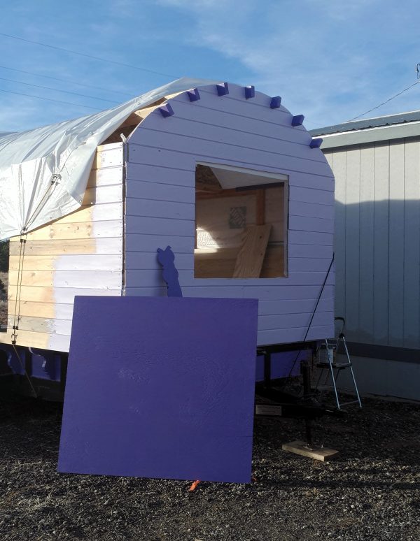 The back wall, now painted the same pale lavender as the front; the short exposed parts of the rafters are the same darker purple as the half-sheet of plywood leaning against the tinker's wagon.