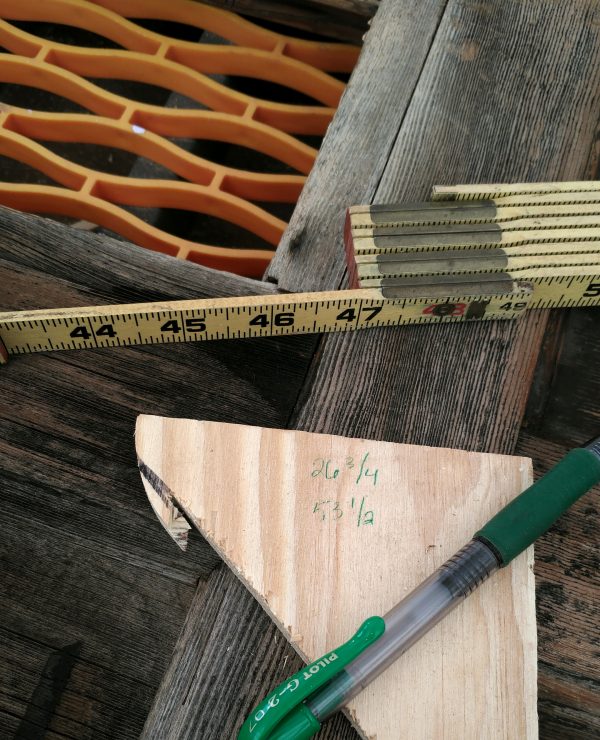 A still life arranged atop the old door: a green pen, a folding ruler, & a scrap of wood with the numbers '26 1/4' & '53 1/2' written upon it in green ink.