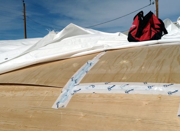 The metal pieces, along with the edge where the plywood overpals, have all been covered in tyvek tape. Also, a red toolbag is sitting on the roof, holding down the white tarp that'll go over everything else.