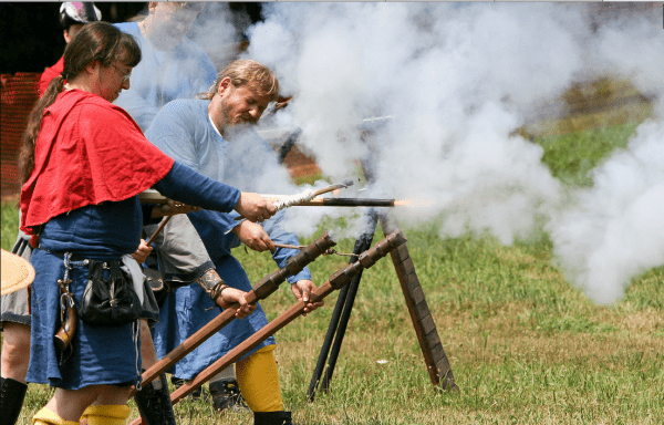 The photo is mostly smoke over green grass, but a couple of people in medieval clothing are visible, along with a couple of, well, small cannons on sticks. The photo is described further in the text.