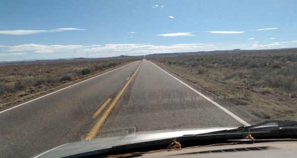 A two-lane road leads straight through flat, scrubby desert under a blue sky, all seen from inside the van.
