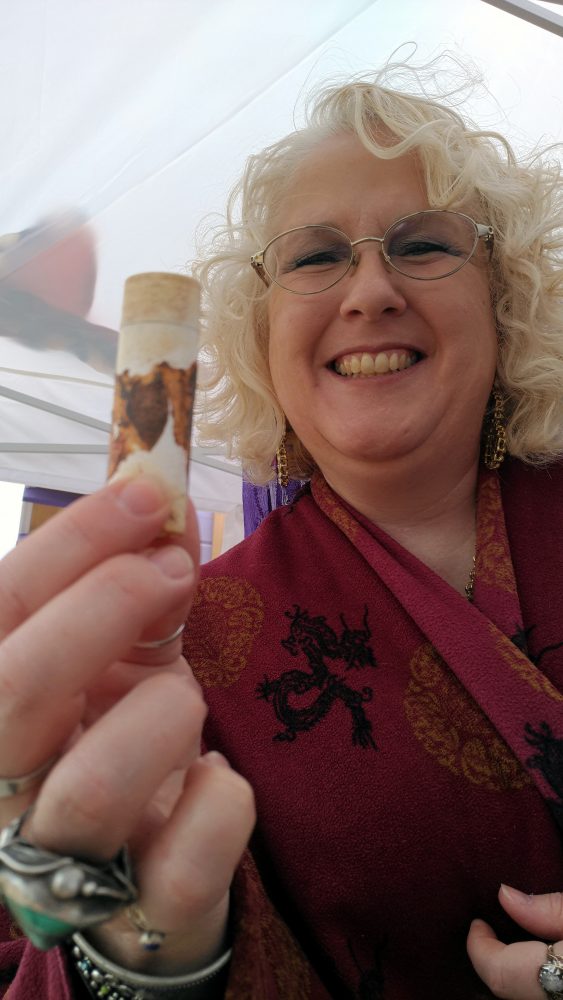 A white lady with curly blond hair, wire-rimmed glasses, & a really glorious smile, holding up a tube of lip balm that has the barest remains of a brown label. This thing has been through a LOT.