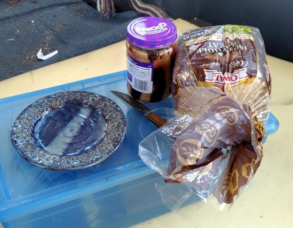 Inside the van, a plate, knife, loaf of bread, & jar of mixed peanut butter & jelly sit on a blue plastic box.