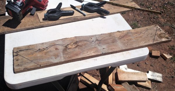 A rough, weathered board lays on a table outside. The same trim pattern that runs up the corners of the tinker's wagon is traced on it in sharpie.