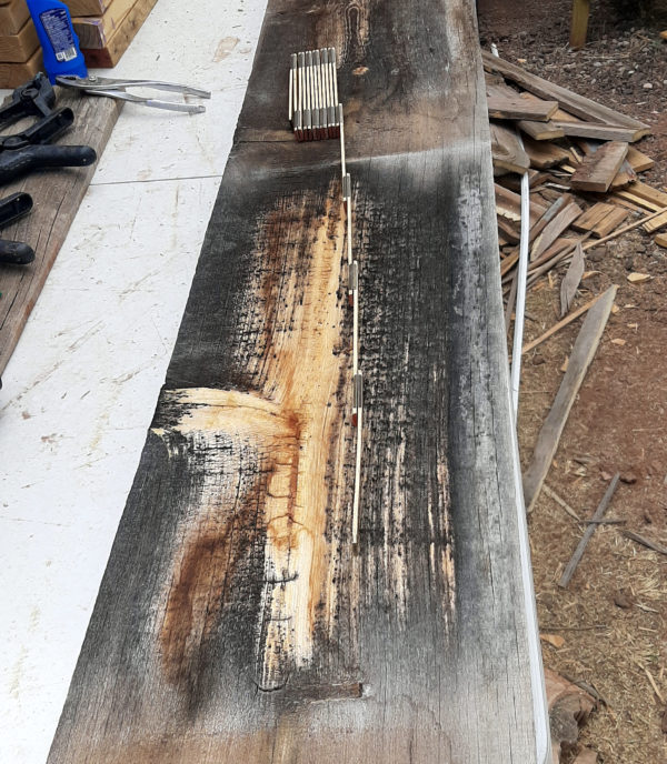 A rough wooden board lays on a table outside, with a folding ruler atop it. Th eboard is about a foot wide & several feet long; most of it is weathered to dark brough, but there's a pale streak down the middle, & a knot on the left side that's also pale.