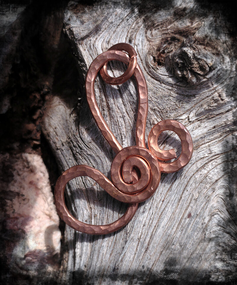 Overlapping spirals in hammered copper are joined by a pair of arching curves, one rising upwards, the other ducking slyly off to the left.