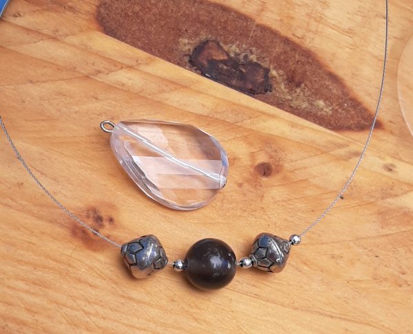 A small faceted glass pendant & a bit of jewelry wire strung with black & silver-coloured beads sit on a wood surface.