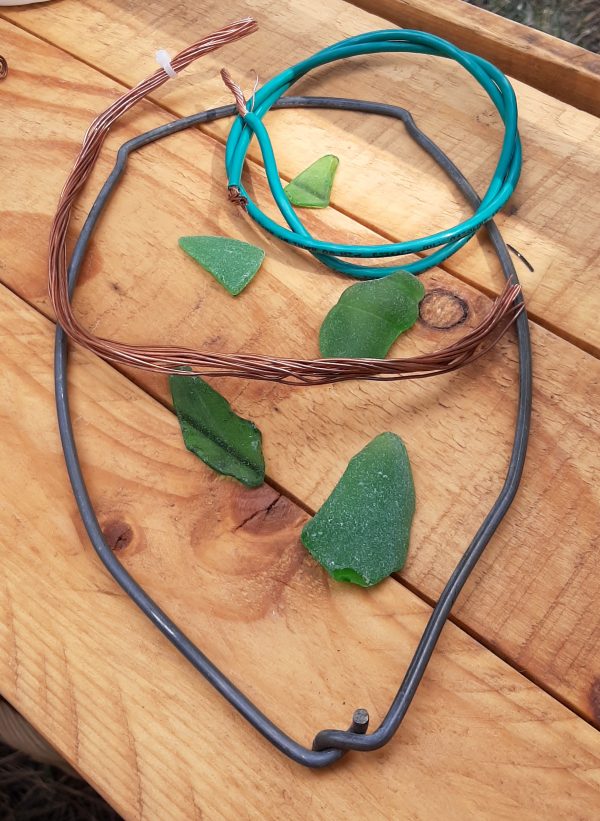 On a wood table: five pieces of green sea glass, a metal bucket handle with the two ends that would go into the top of the bucket twined together, & two buncldes of different thicknesses of copper wire, one enclosed in a teal-blue coating.