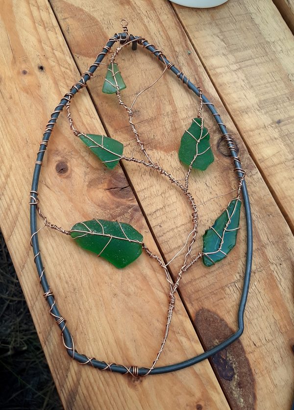 The mostly-finished piece. The bucket handle acts as a frame; it's broad at the bottom and narrows to a point at the top where the two ends are twined together. A long vine of copper winds its way up the middle, with five green sea glass leaves, wrapped in copper, emerging from it.
