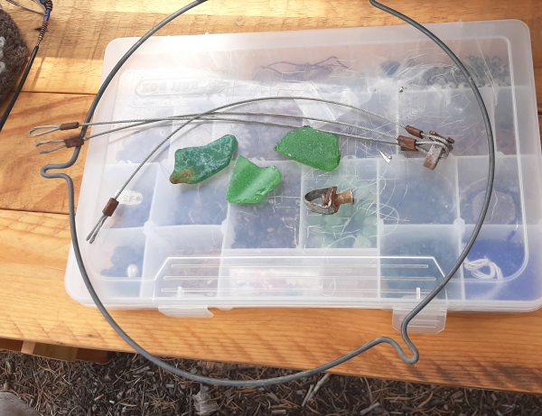 On top of a bead organizer: three pieces of green seaglass, a more-or-less round metal hoop with three equally-spaced u-shaped bumps to the outside, & three lengths of braided steel wire, joined together at the ends by a triangular bit of metal.