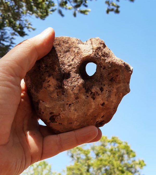 My hand holding a rock up to the sky. It's perhaps the size of my fist, mottled in shades of brown & reddish tan, & the sky is visible through a pinky-sized hole.