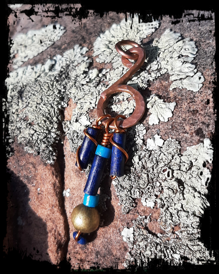 Thick copper wire bent into the shape of a music note. Three strands of beads, in dark blue, teal, and brass, hang from the bottom curve.