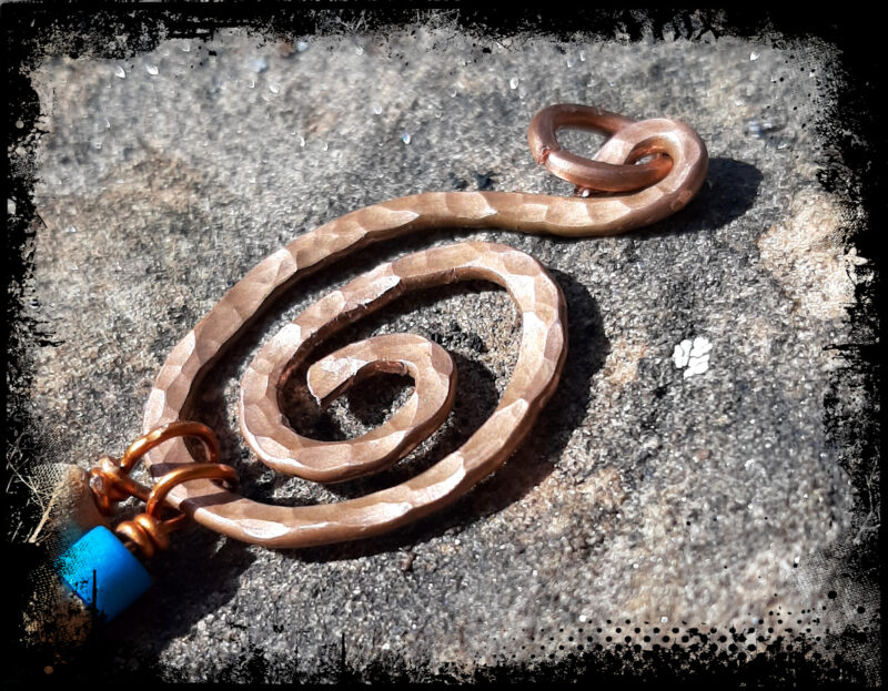 A spiral of hammered copper wire. Two lengths of thinner copper wire, threaded with blue and tan wooden beads, hang from the bottom.