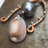 A tall, uneven arc of copper wire arches over and around a strand of silver-colored wire threaded with a big dark grey bead. A cowrie shell hangs below the bead.