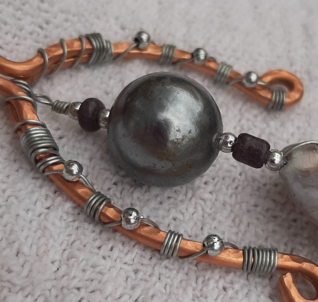A tall, uneven arc of copper wire arches over and around a strand of silver-colored wire threaded with a big dark grey bead. A cowrie shell hangs below the bead.