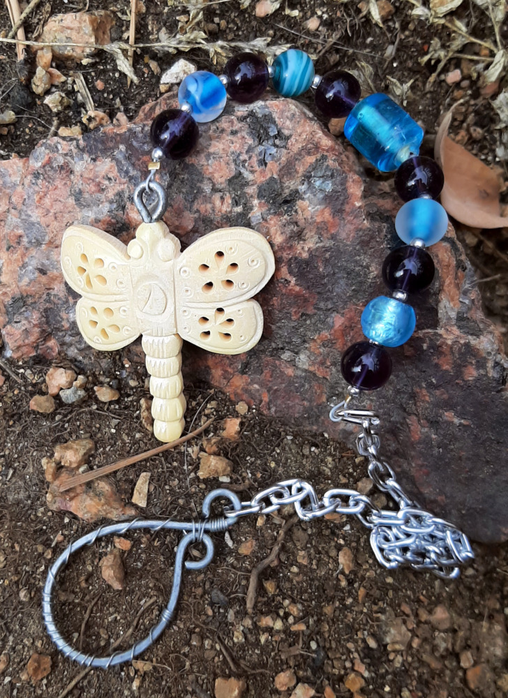 A pendulum with a dragonfly carved out of what looks like bone at one end, then glass beads alternating teal and purple, then a length of silver-colored chain, and topped by a loop of steel wire hammered flat with much thinner silver-colored wire wrapped around it.