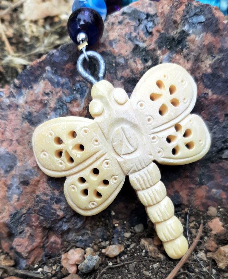 A close view of the dragonfly. It's roughly carved into something the pale yellowish color of bone. It's pretty stylized, with five small holes in each wing, arranged like the petals of a flower.