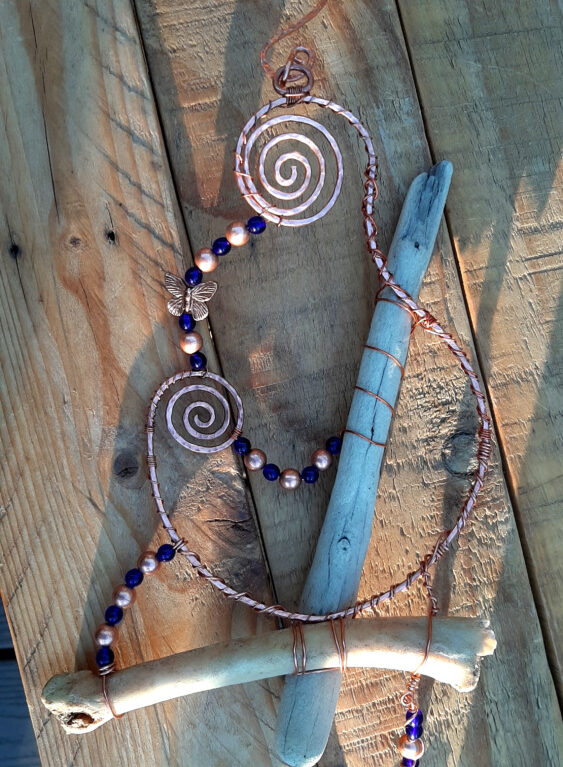 Almost-vertical driftwood meets a gently arched leg bone. Overall is a long sweep of curved copper, spiralled at each end. Strands of blue and copper beads festoon, arching between the larger elements.