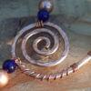 A hammered copper spiral wound with thinner wire. Strands of beads sprout from it, one up, one down and to the left.