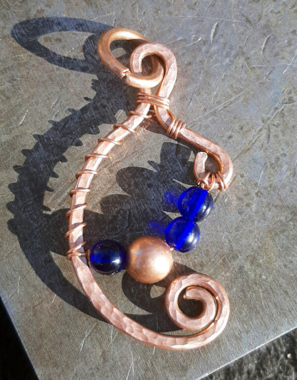 Copper wire in a rough C shape with a loop at the top and a spiral at each end. Thinner copper wire holds an arc of beads across -- blue, copper, blue, blue.