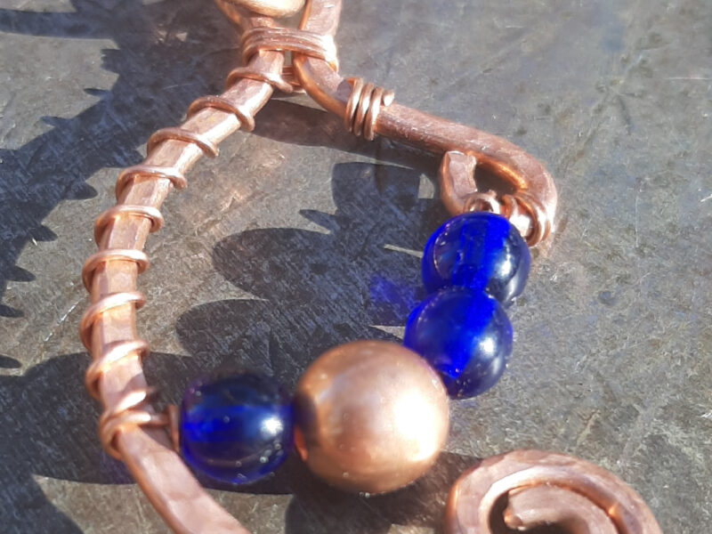Copper wire in a rough C shape with a loop at the top and a spiral at each end. Thinner copper wire holds an arc of beads across -- blue, copper, blue, blue.