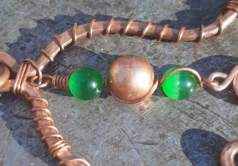 An asymmetrical arch in copper wire -- one end turned inwards, the other curved out. Hanging down the middle are two green glass beads with a copper one between them, all threaded onto thin copper wire.