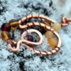 A curving, looping shape in copper wire is adorned with round glass beads ranging in color from yellow through various ambers to red.