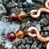 Each earring curves hammered copper down into and through a single loop with a pale yellow glass bead; two more, amber then deep red, hang below.