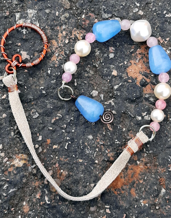 A pendulum; the bottom is a vaguely cone-shaped pale blue bead threaded onto silver-colored wire bent into a spiral shape at the bottom. Above that are alternating pink and white beads, interspersed with two more of the blue ones. A length of narrow white leather leads to a round loop of bright copper wire decoratively wrapped in much narrower copper wire.